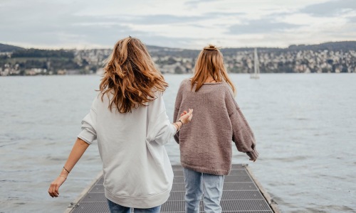 two women walk to the edge of a dock, in a body of water, looking toward the city on the other shore